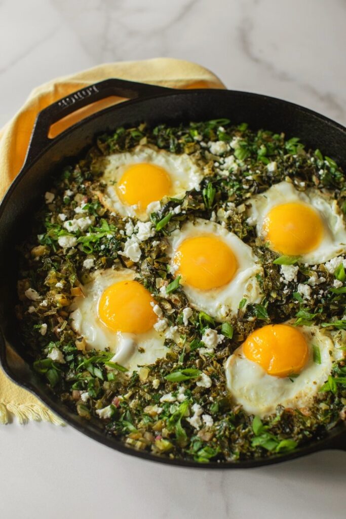 Bright green shakshuka cooked in a black skillet with eggs nestled into wilted kale, spinach, and Brussels sprouts, topped with feta and herbs.”