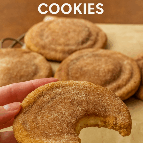 A close-up of a hand holding a high-protein churro cheesecake cookie with a bite taken out, revealing the creamy cheesecake filling inside. The cookie is coated in cinnamon sugar and surrounded by more cookies in the background, all resting on parchment paper.