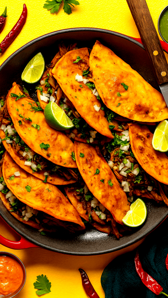 Birria tacos served with dipping consomé in a ceramic pot on a wooden counter.