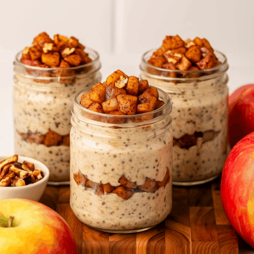 Four glass jars filled with creamy high-protein apple pie overnight oats topped with fresh apple slices, cinnamon, and a sprinkle of granola, styled on a rustic wooden table.