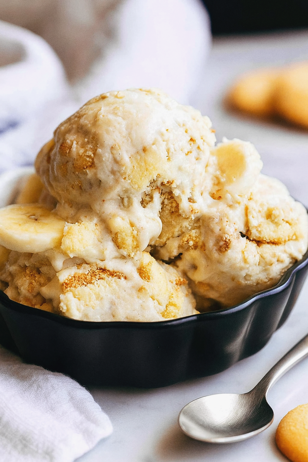 Low sugar banana pudding in a black bowl on a white marble surface with a silver spoon and white towel, topped with banana slices and cookie crumbles
