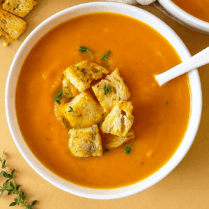 Creamy butternut squash soup served in a white bowl with a white soup spoon, topped with golden croutons and thyme on a warm rustic table, high-protein dairy-free fall recipe.