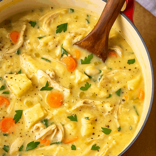 Cozy homemade chicken and dumpling soup simmering in a red Dutch oven with a wooden spoon and grey kitchen towel on the side, creamy broth filled with tender chicken, carrots