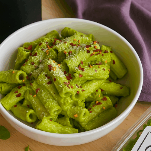 A white ceramic bowl filled with creamy super green high protein pasta made with chickpea pasta, spinach, and Greek yogurt sauce, styled on a wooden table with a soft napkin and a black cup beside it.