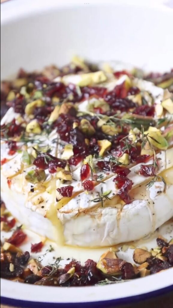 Warm baked brie topped with pistachios, cranberries, honey, and thyme on a rustic wooden board — the perfect festive Christmas appetizer.