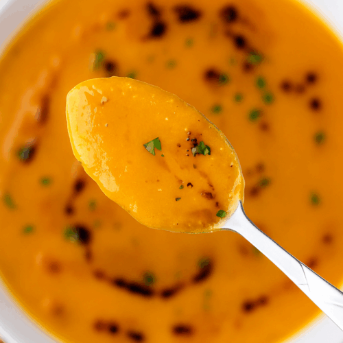 A warm bowl of creamy high protein Turkish lentil soup made with red lentils, carrots, and spices, topped with a drizzle of olive oil and fresh herbs, served in a white bowl on a cozy kitchen table