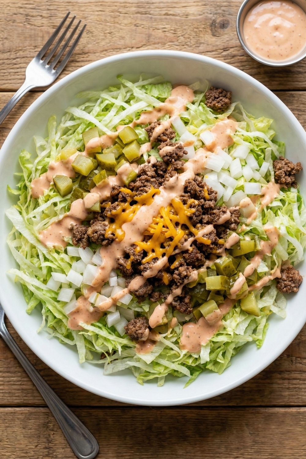 Top-down view of a large low-carb Big Mac salad bowl featuring shredded iceberg lettuce, seasoned ground beef, diced pickles, white onions, and a drizzle of sugar-free special sauce.
