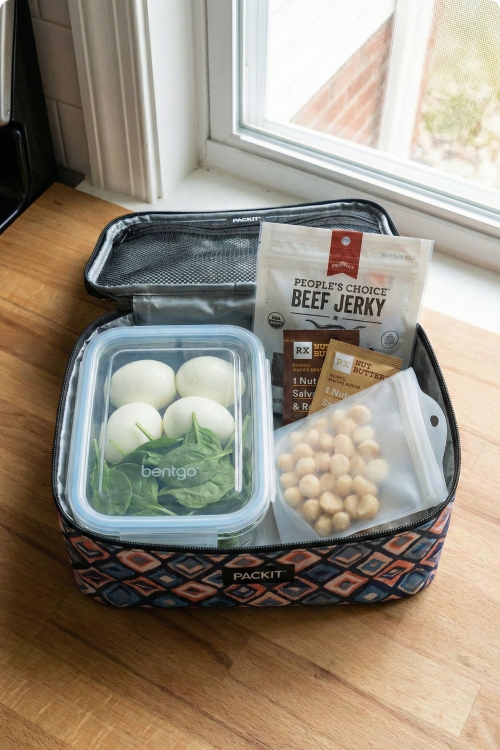 Low carb travel snack prep on kitchen counter featuring PackIt cooler bag, Bentgo box with boiled eggs, and sugar-free beef jerky.