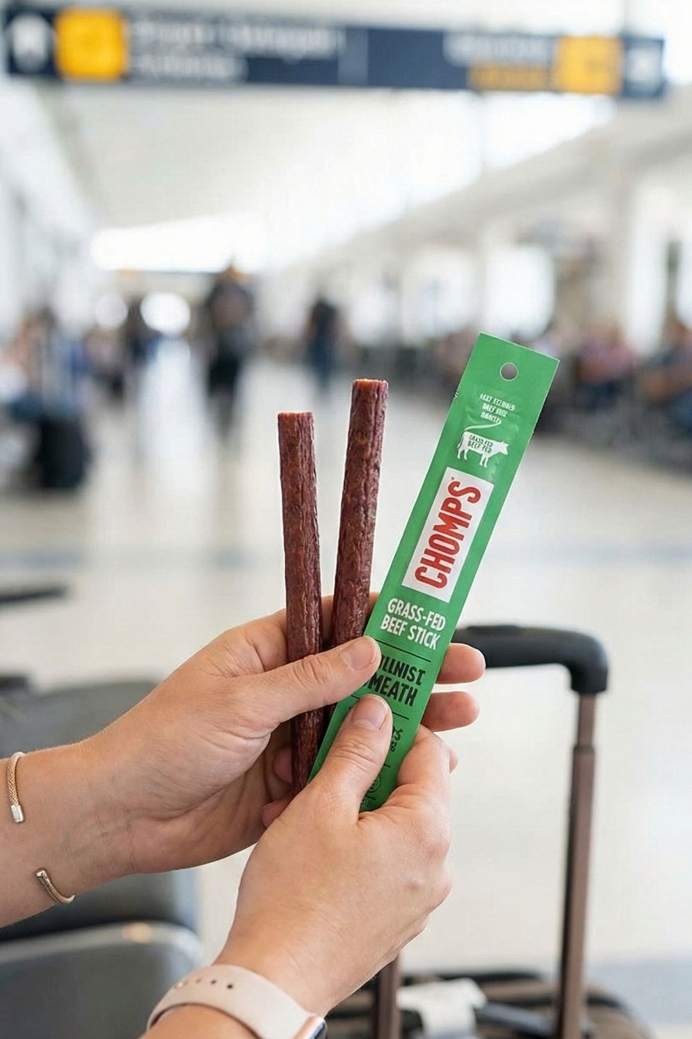 Woman holding a Chomps Grass-Fed Beef Stick at an airport terminal. A mess-free, TSA-approved high-protein snack for healthy travel.