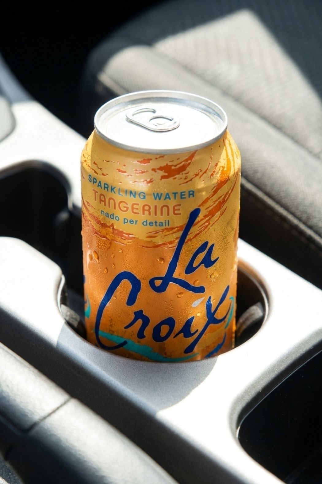 Cold can of Tangerine LaCroix sparkling water sitting in a sunlit car cup holder for sugar free hydration on the road.