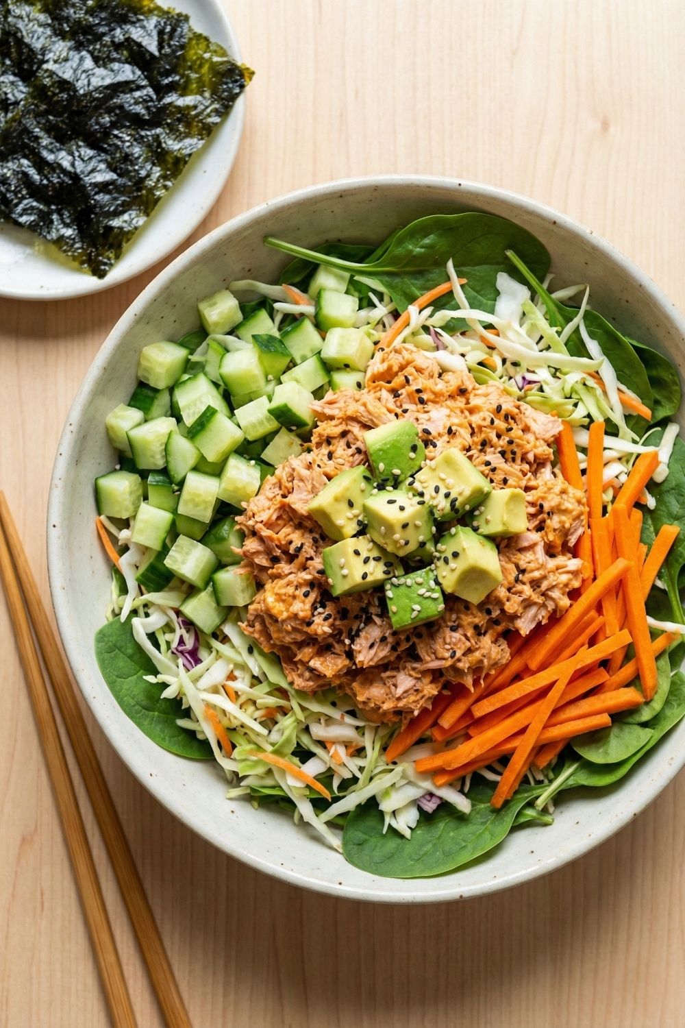 Overhead shot of a high-volume spicy tuna salad bowl with fresh cabbage, cucumber slices, chunks of tuna, avocado, and a spicy mayo dressing, styled like a deconstructed sushi roll.