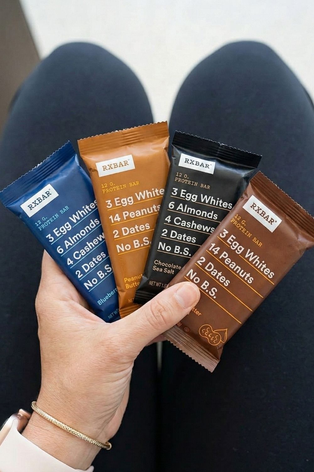 Hand holding a variety of RXBAR clean ingredient protein bars at an airport gate. The best whole-food emergency snack for long flights.