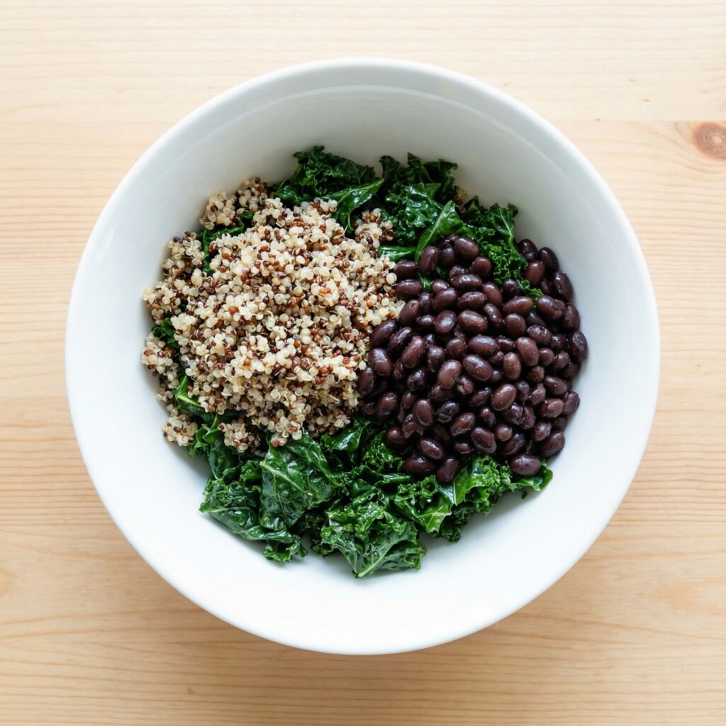 Assembling a healthy lunch bowl with a base of massaged kale, featuring sections of cooked quinoa and black beans arranged neatly in a white bowl.