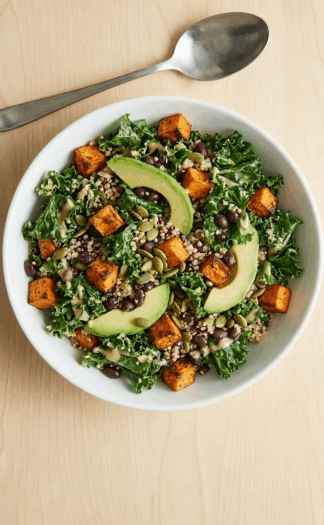 Wide overhead shot of a fully mixed roasted sweet potato and kale power bowl in a white ceramic bowl, ready to eat, with a serving spoon resting beside it on a wooden surface