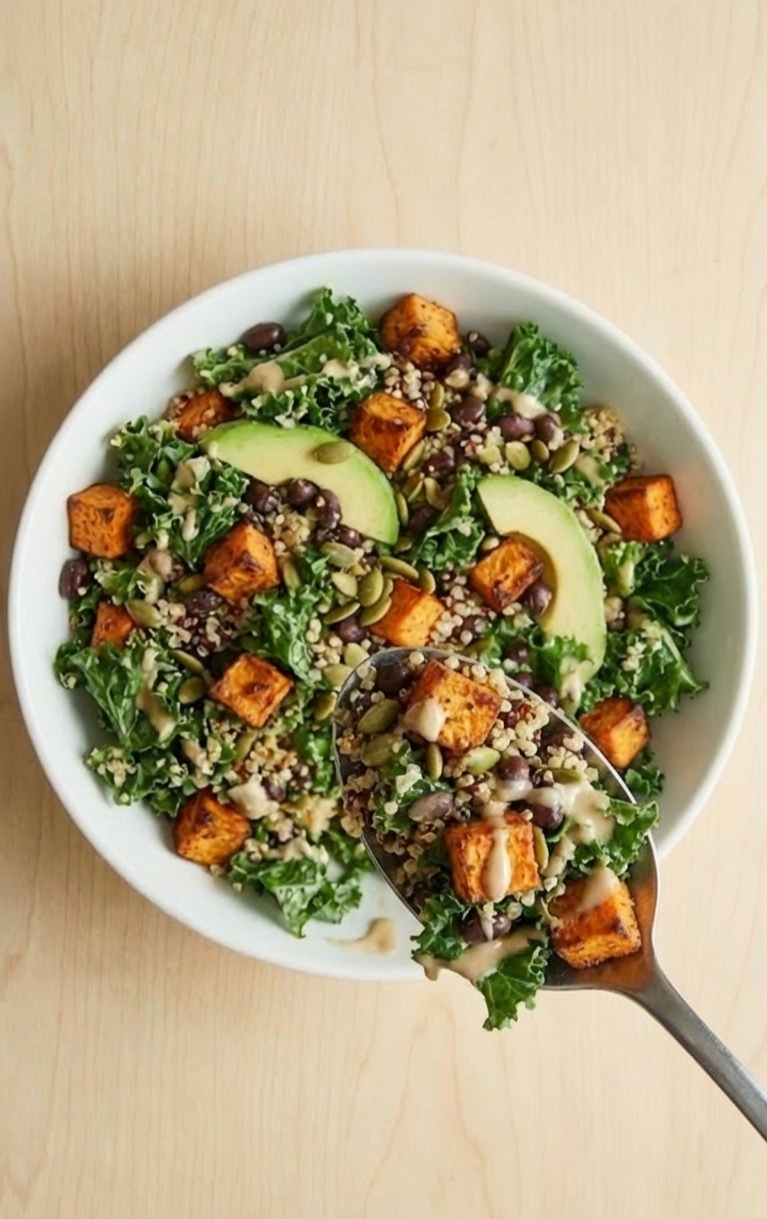Overhead shot of a fully mixed roasted sweet potato and quinoa salad in a speckled ceramic bowl, with a spoon lifting a generous serving of the vegan power bowl