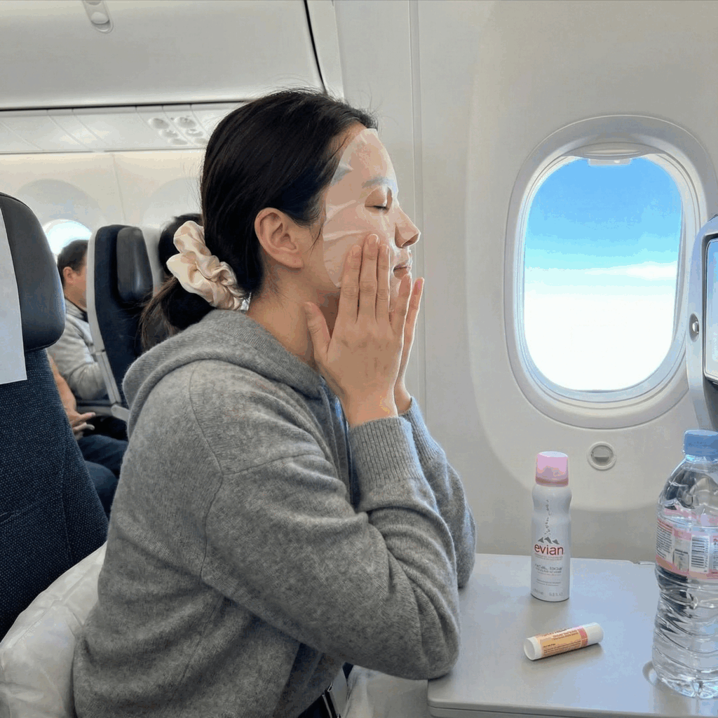 Woman relaxing in an airplane window seat wearing a hydrating sheet mask and cozy hoodie, demonstrating a proper in-flight skincare routine for long-haul travel.