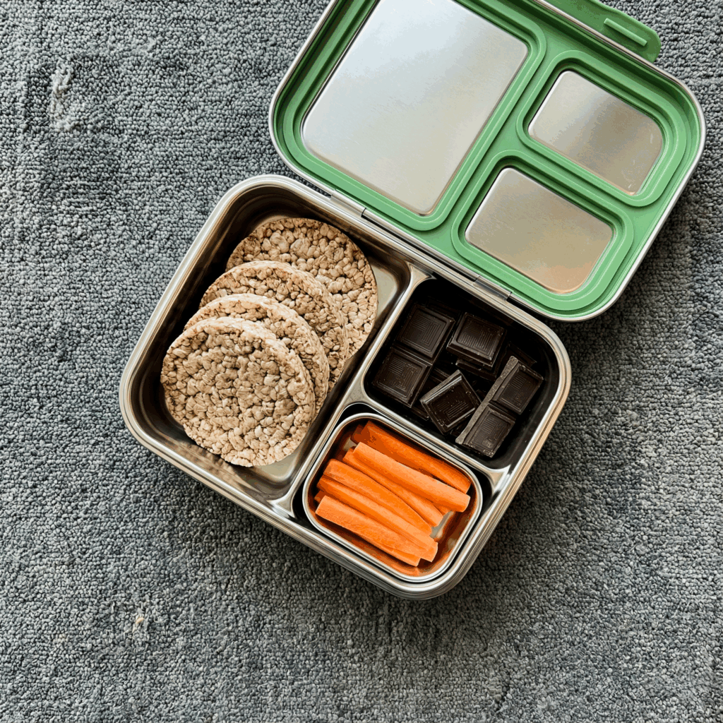 Top-down view of an open stainless steel bento box packed with brown rice cakes, dark chocolate squares, and carrot sticks sitting on airport gate carpet, showing organized and gluten-free healthy travel snacks.