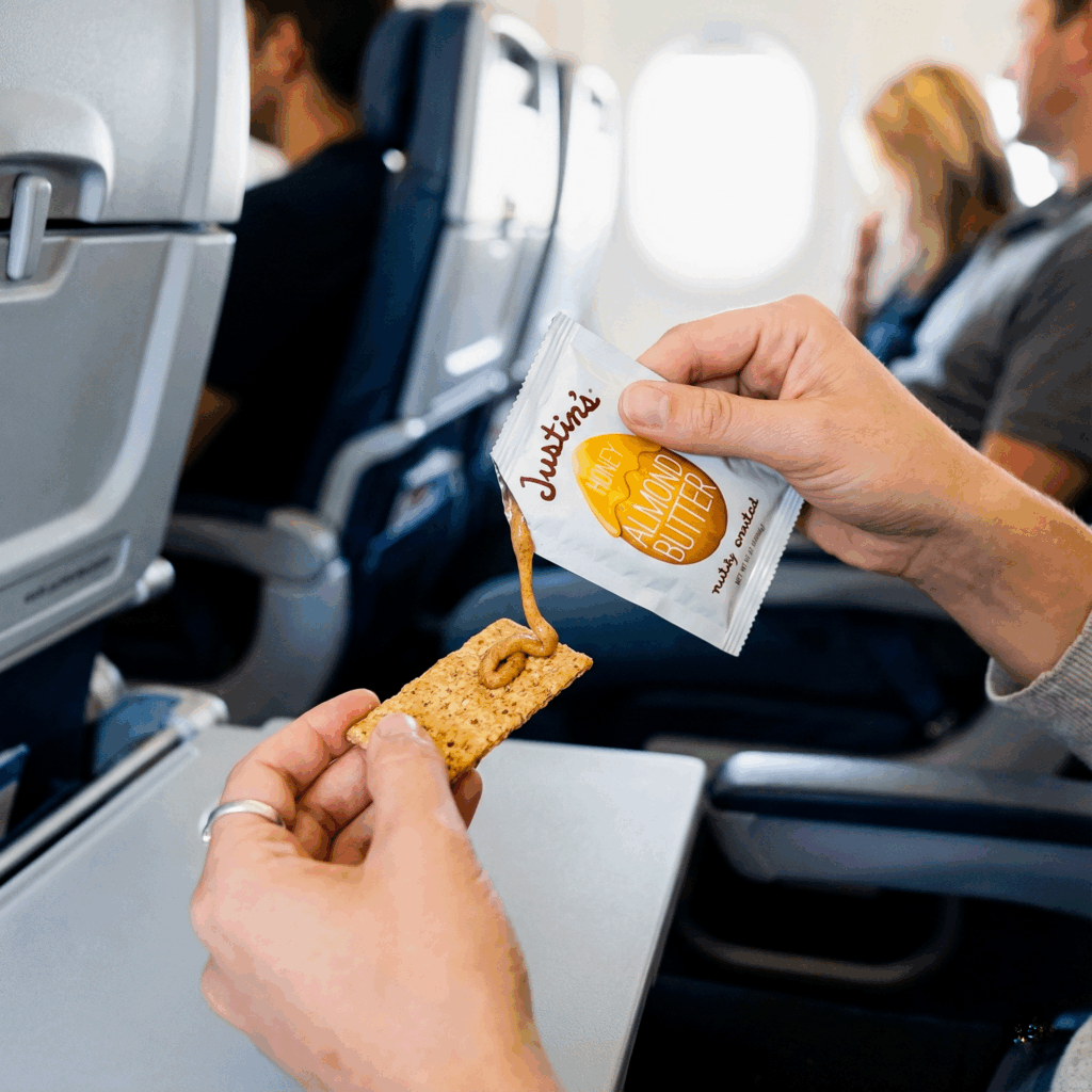 Close-up of a woman's hands squeezing a single-serve almond butter packet onto a whole-grain cracker while seated on an airplane, demonstrating a high-protein, mess-free travel snack idea.