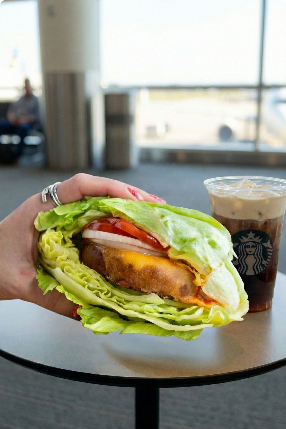 Healthy airport food option showing a lettuce-wrapped burger and iced coffee with heavy cream held by a traveler.