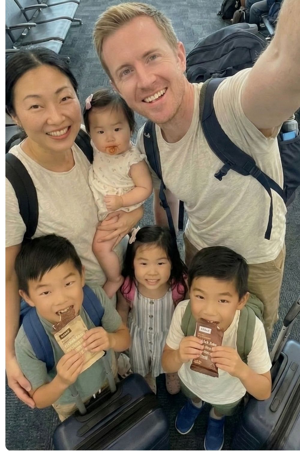 A high-angle selfie of a family of six at a busy airport terminal gate before a flight. The father takes the photo while the mother holds a baby with a messy face, and two young sons are eating chocolate protein bars amidst their carry-on luggage.