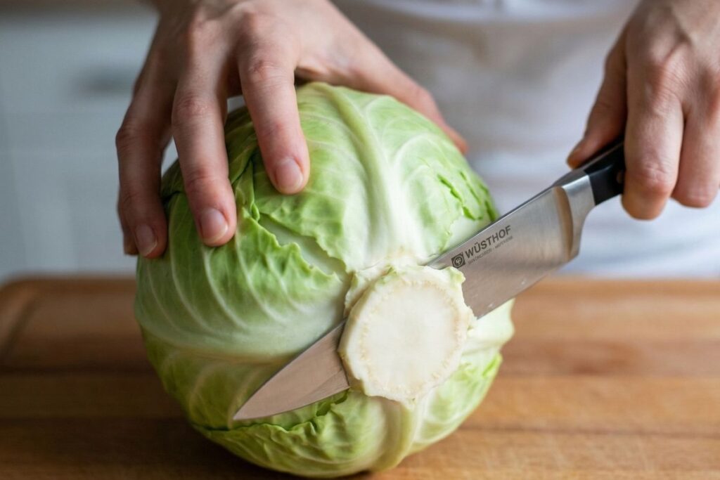 Woman's hands using a Wüsthof chef's knife to slice through the white core of a green cabbage, keeping the stem attached so the steaks don't fall apart.