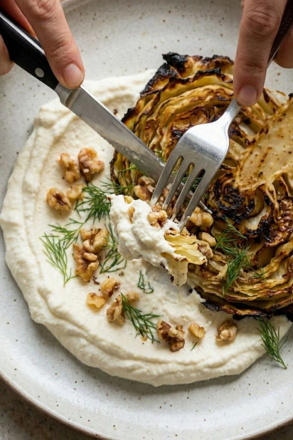 Close-up of a fork cutting into a tender roasted cabbage steak with creamy whipped feta and walnuts, showing the soft texture and crispy edges