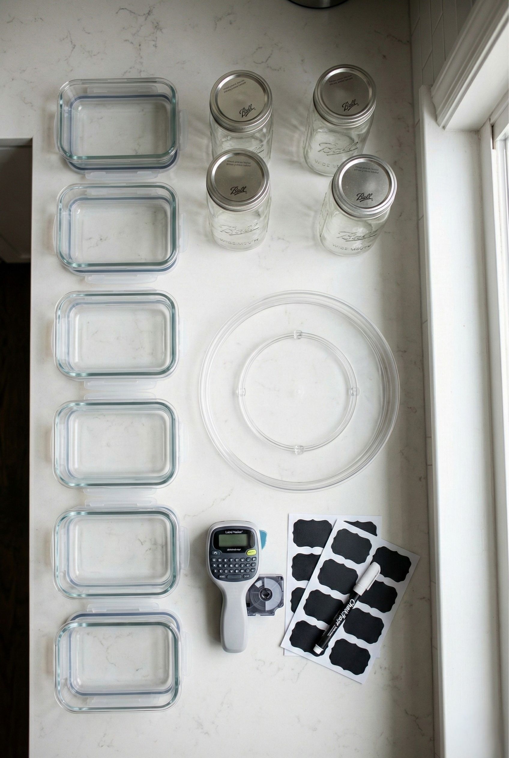 Kitchen counter display of essential meal prep gear, including empty glass storage containers, wide-mouth mason jars, chalkboard labels, and a clear turntable.