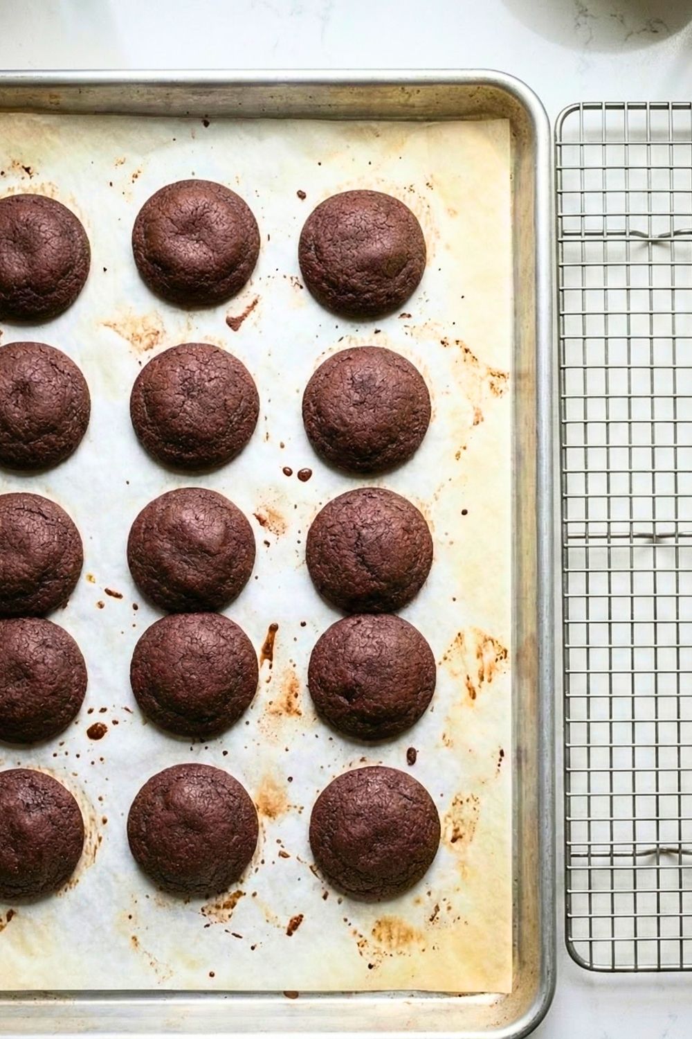 Overhead view of freshly baked black cocoa cookies on a baking sheet with parchment paper next to a wire cooling rack.