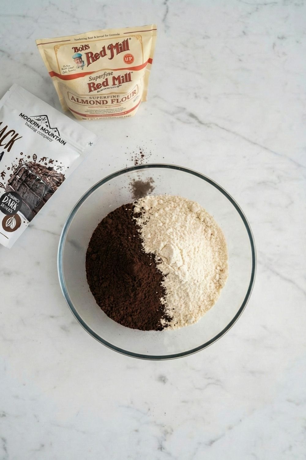 Overhead flat lay of a clear glass bowl containing white superfine almond flour and jet-black cocoa powder for a homemade zero sugar Oreo recipe.