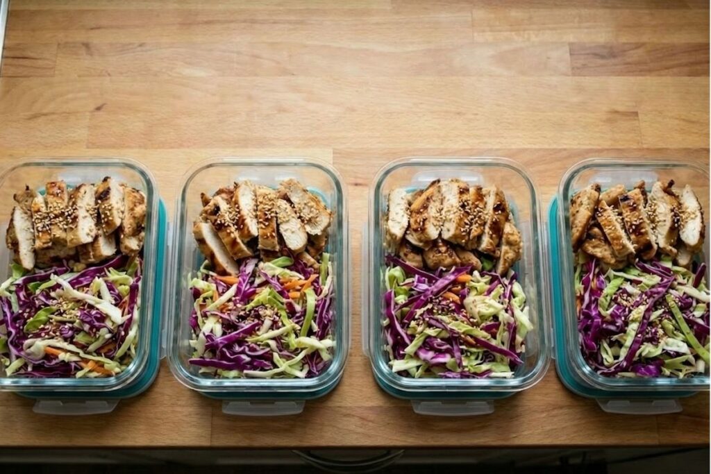 Four Pyrex glass meal prep containers stacked and arranged containing high protein cabbage salad and grilled chicken.
