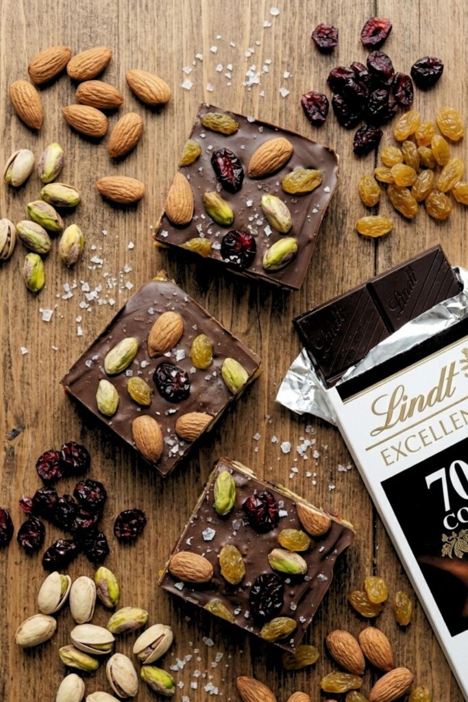 Homemade fruit and nut chocolate bars displayed on a marble board next to raw ingredients and a Lindt 70% cocoa bar.