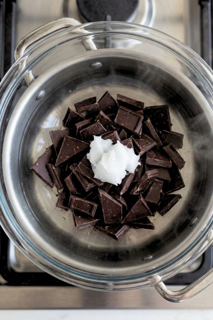 A heat-proof bowl placed over a pot of simmering water containing broken dark chocolate pieces and coconut oil