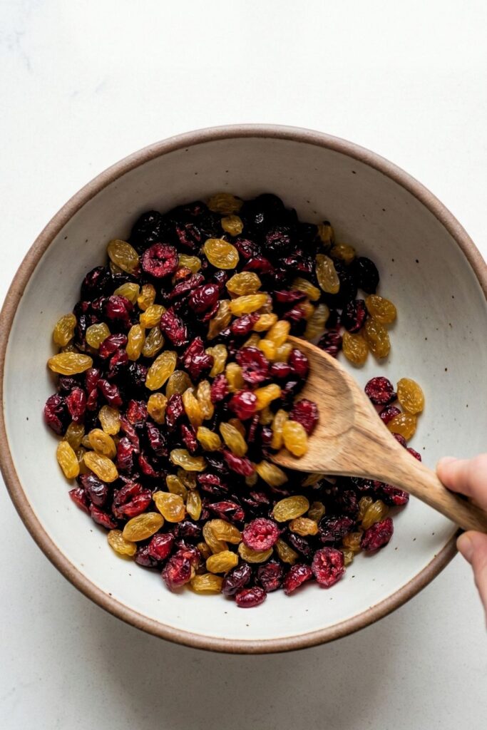 A bowl filled with raw almonds, pistachios, dried cranberries, and golden raisins being mixed together