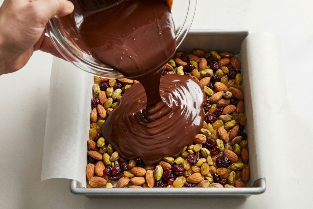 Pouring melted dark chocolate over a base layer of nuts and fruit in a square baking pan.