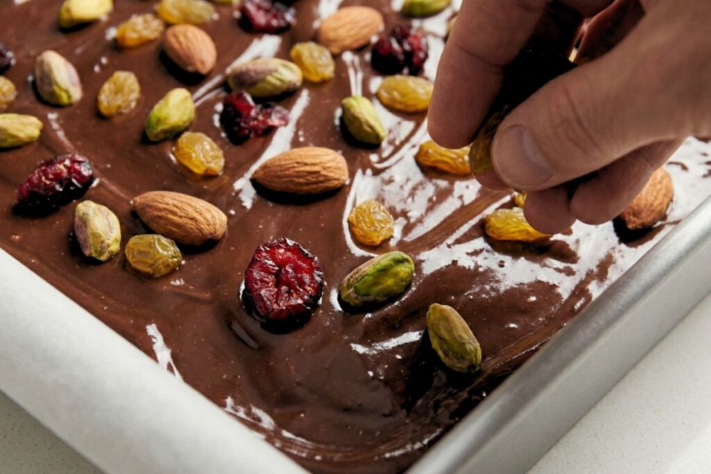Sprinkling remaining pistachios, cranberries, and raisins on top of wet dark chocolate for decoration.