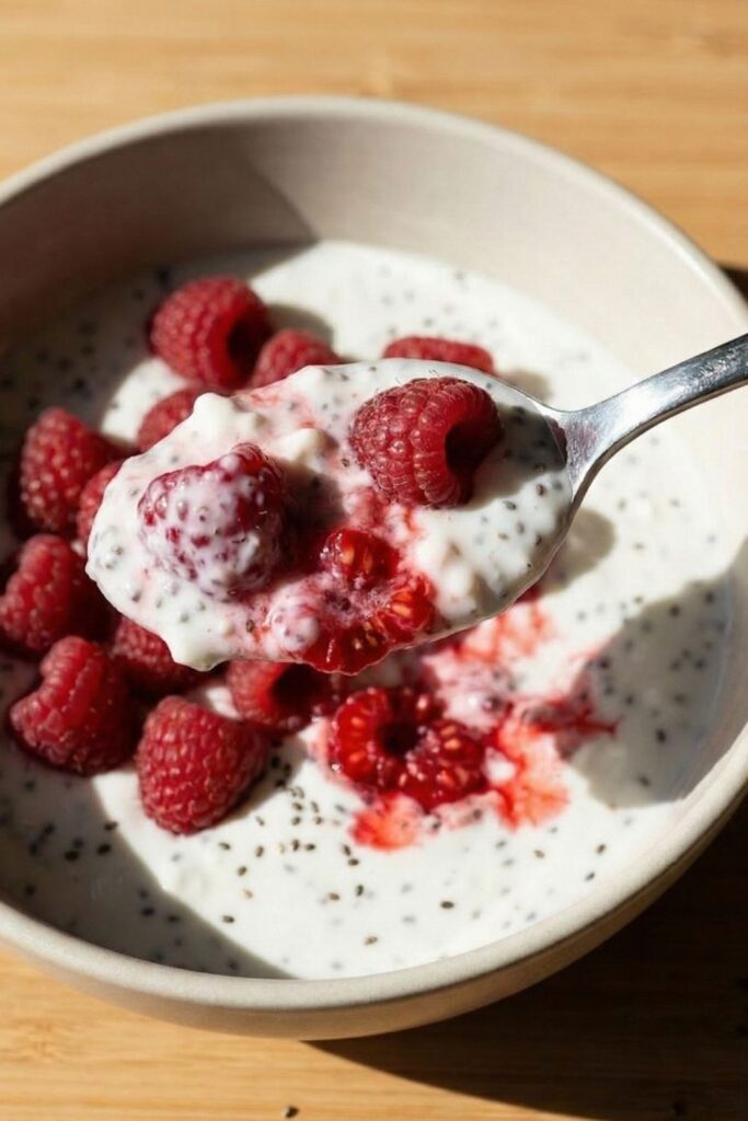 A close-up spoonful of creamy cottage cheese and chia seed pudding topped with a raspberry, showing the thick, luscious texture and overhead lighting.