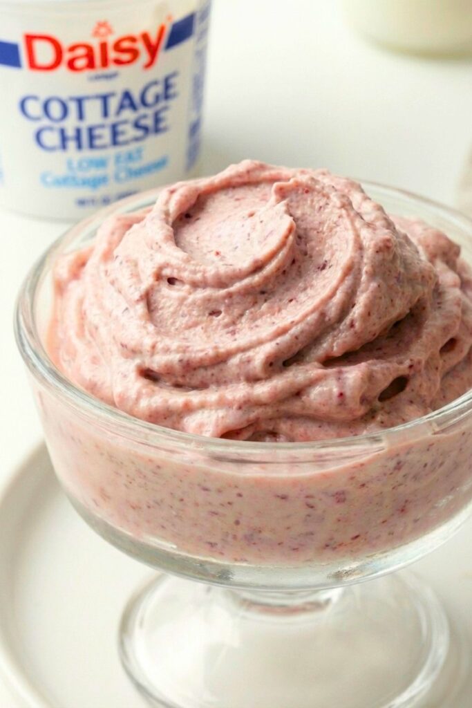 A large bowl of fluffy, creamy high protein ice cream swirled like soft serve, with a blurry cottage cheese container in the background.