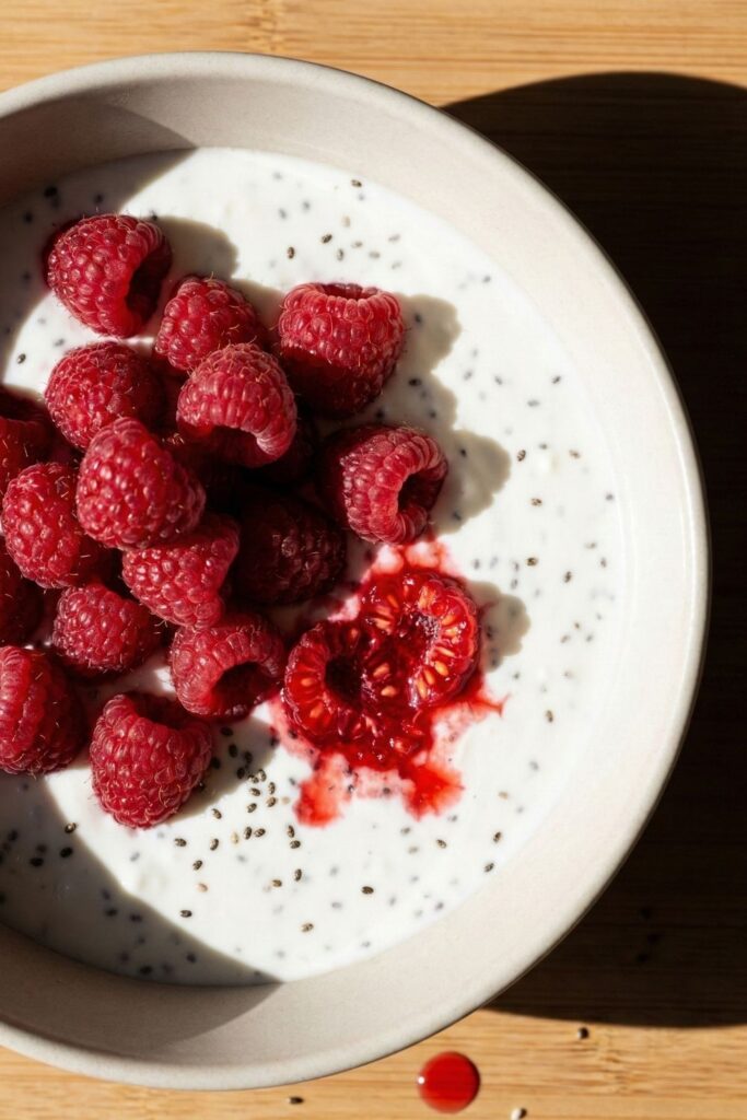 A finished high-protein, GLP-1 friendly breakfast bowl filled with whipped cottage cheese and chia seeds, topped with a pile of fresh raspberries and red juice drips in hard light.