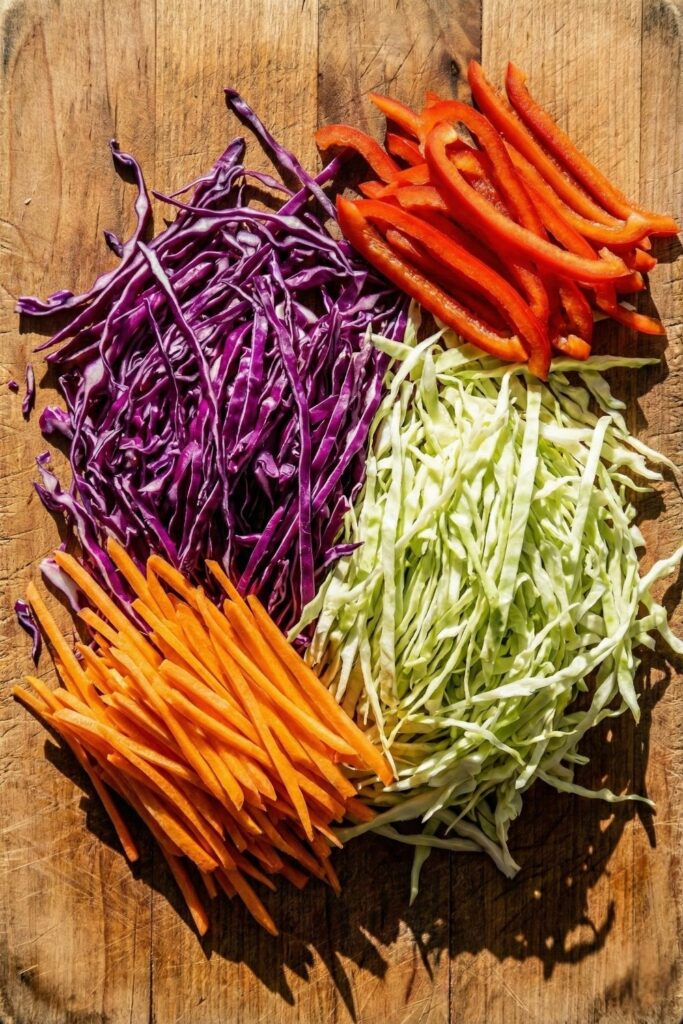 Vibrant salad ingredients arranged on a cutting board, including piles of purple cabbage, green cabbage, julienned carrots, and sliced red bell peppers, displaying a colorful high-fiber mix.