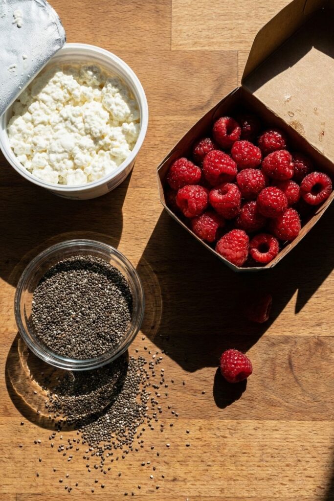 Overhead shot of healthy breakfast ingredients on a wooden table, featuring an open tub of cottage cheese, a glass bowl of chia seeds, and a carton of fresh raspberries under sharp sunlight.