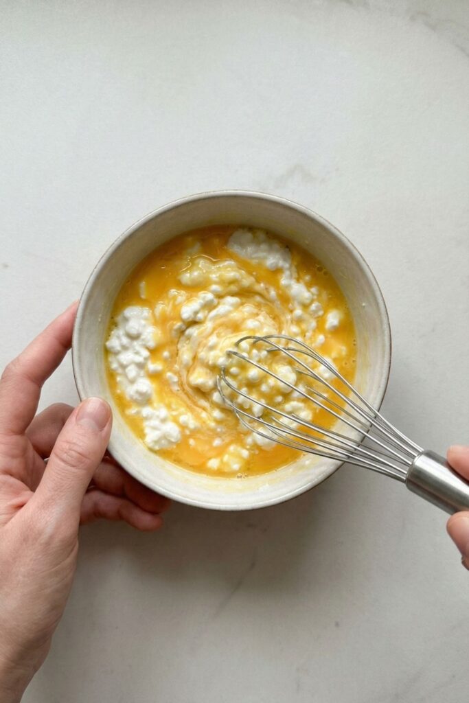 Overhead shot of whisking eggs and cottage cheese in a bowl, showing visible white curds for a high-protein breakfast base.