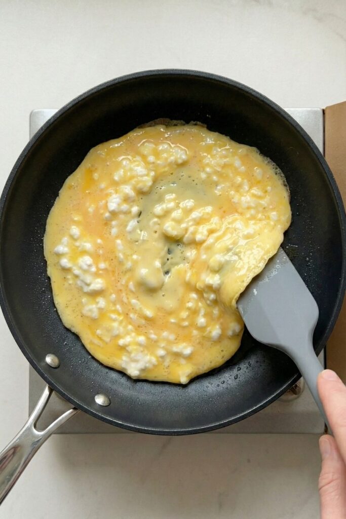 A silicone spatula gently pushing eggs across the pan to form soft, wet curds for the perfect creamy texture.