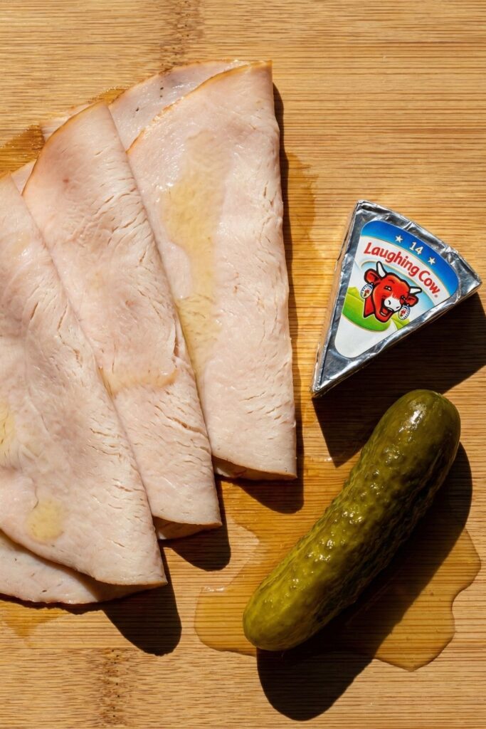 Overhead shot of healthy snack ingredients including deli turkey slices, a wedge of Laughing Cow cheese, and a dill pickle spear with brine on a wooden board.