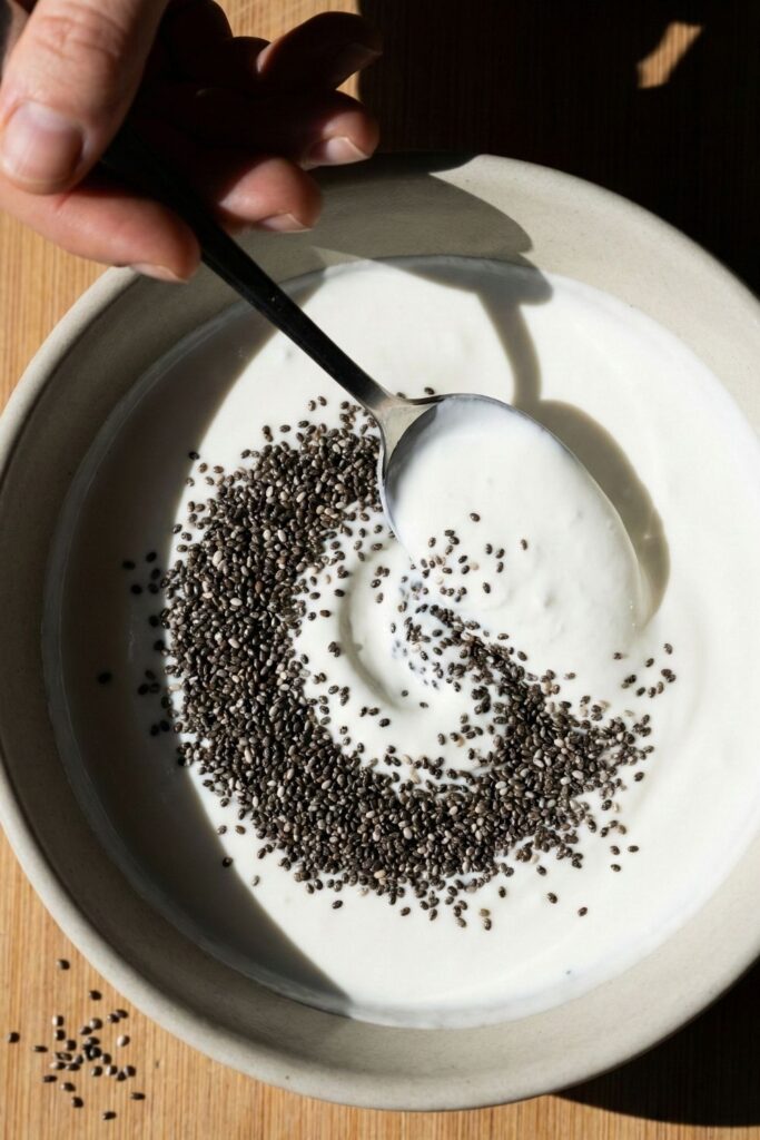 A hand stirring black chia seeds into creamy white whipped cottage cheese in a ceramic bowl, creating a high-contrast swirl pattern.