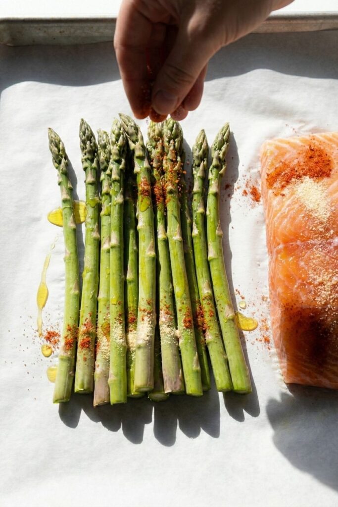 Close-up overhead view of a hand sprinkling paprika and garlic powder seasoning over a raw salmon fillet, showing red spice texture on the fish.