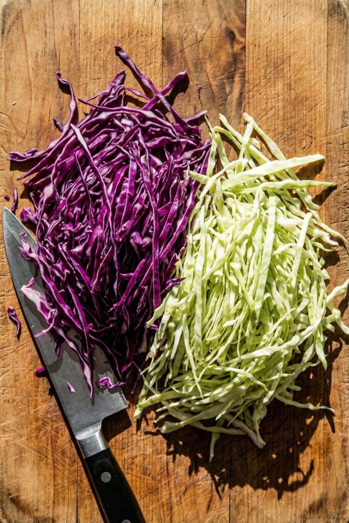 Overhead view of thinly shredded purple and green cabbage on a wooden cutting board with a sharp knife, showing paper-thin texture for a crunchy salad base.