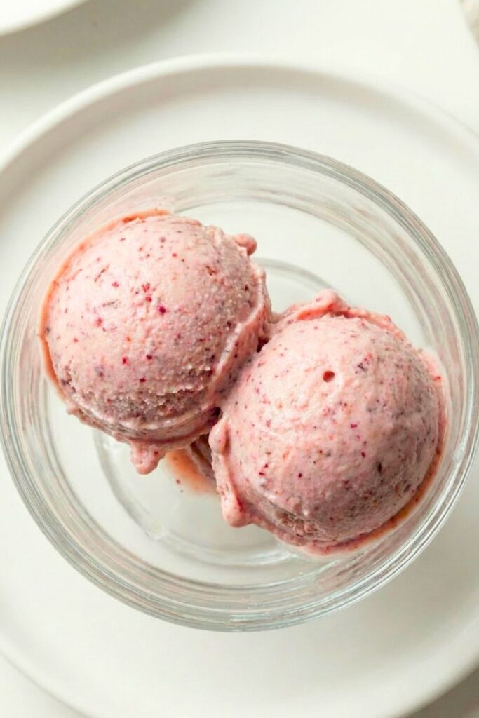 An ice cream scoop forming a perfect round ball of vanilla high protein ice cream from a glass bowl.