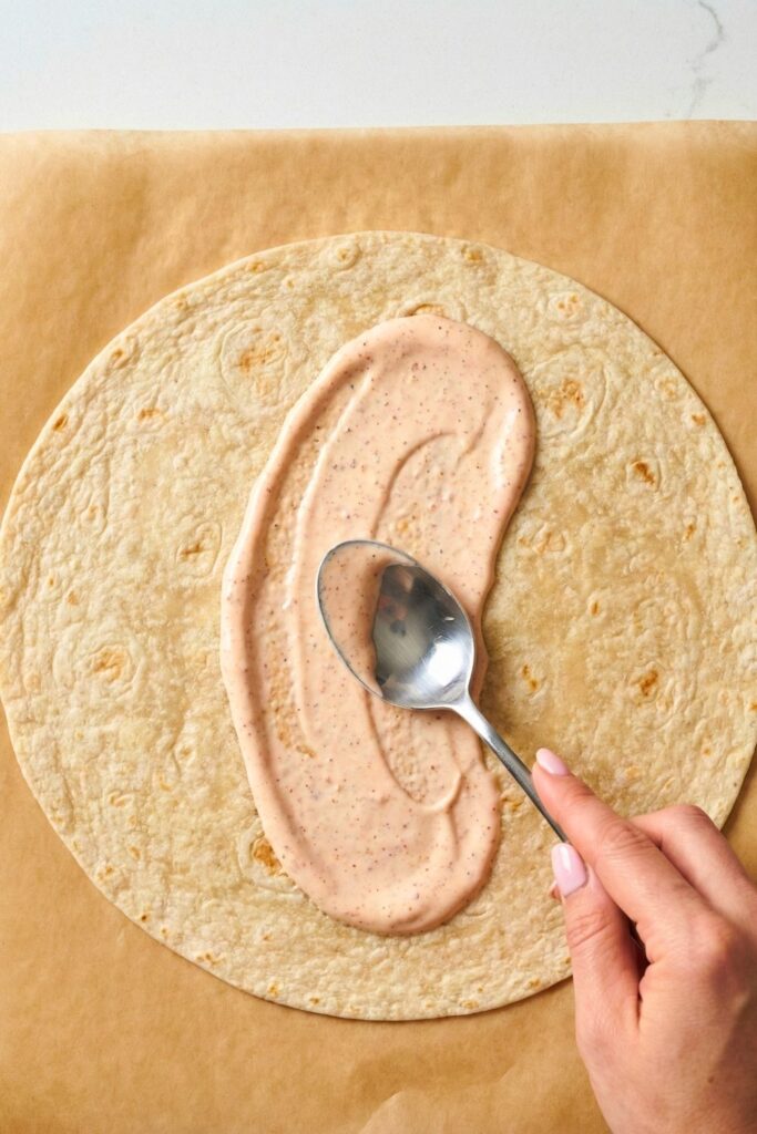 Spreading creamy pink cottage cheese sauce onto a high-fiber tortilla wrap using the back of a spoon.