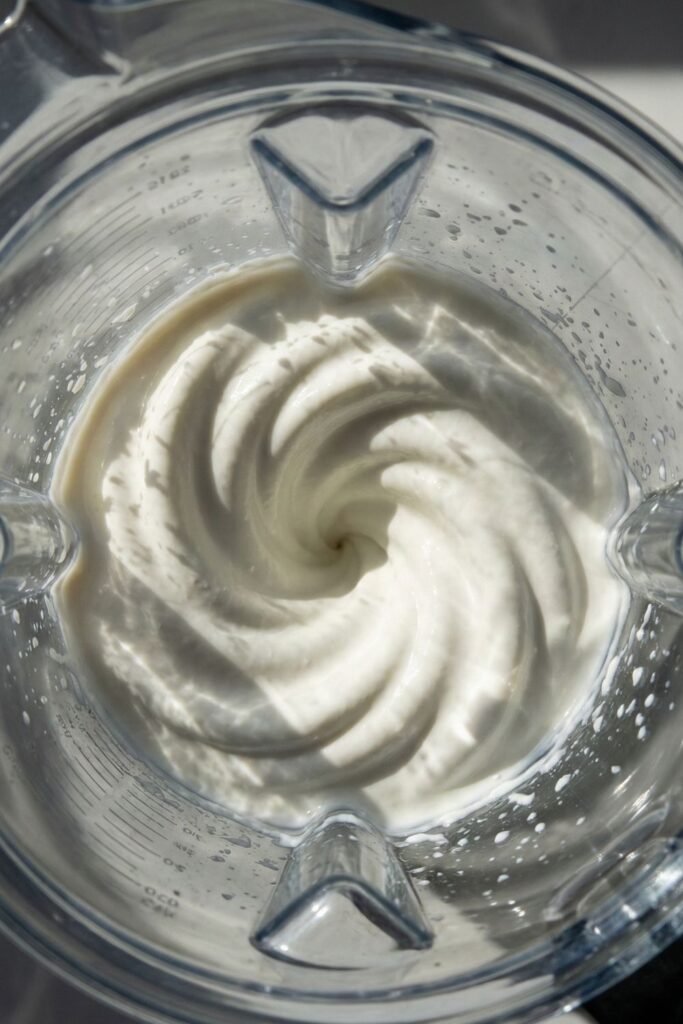 Top-down view into a blender container showing the smooth, glossy texture of whipped cottage cheese with swirl patterns from the blades.