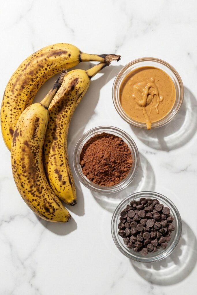 Pre-measured ingredients for healthy 3 ingredient brownies including 3 ripe, spotted bananas, 1/2 cup peanut butter, and 1/3 cup cocoa powder in glass prep bowls on a white marble surface.